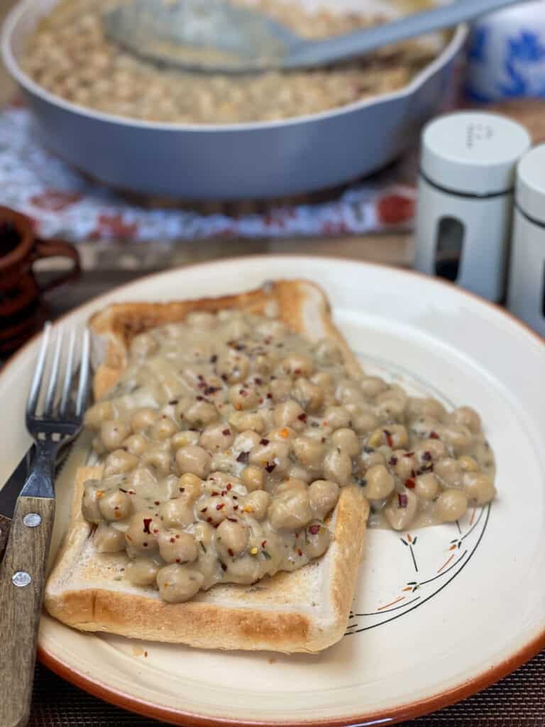 creamed chickpeas scooped over the toasted bread on plate, fry pan to background with grey coloured ladle.