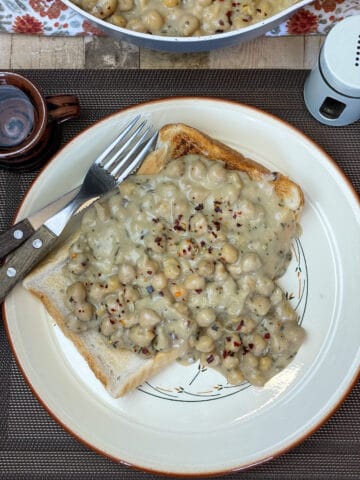 plate of creamed chickpeas on toasted bread with wooden handle fork and knife to side, brown mat and brown small cup, grey pepper pot to side and small glimpse of cooking pan with chickpeas. Featured image.