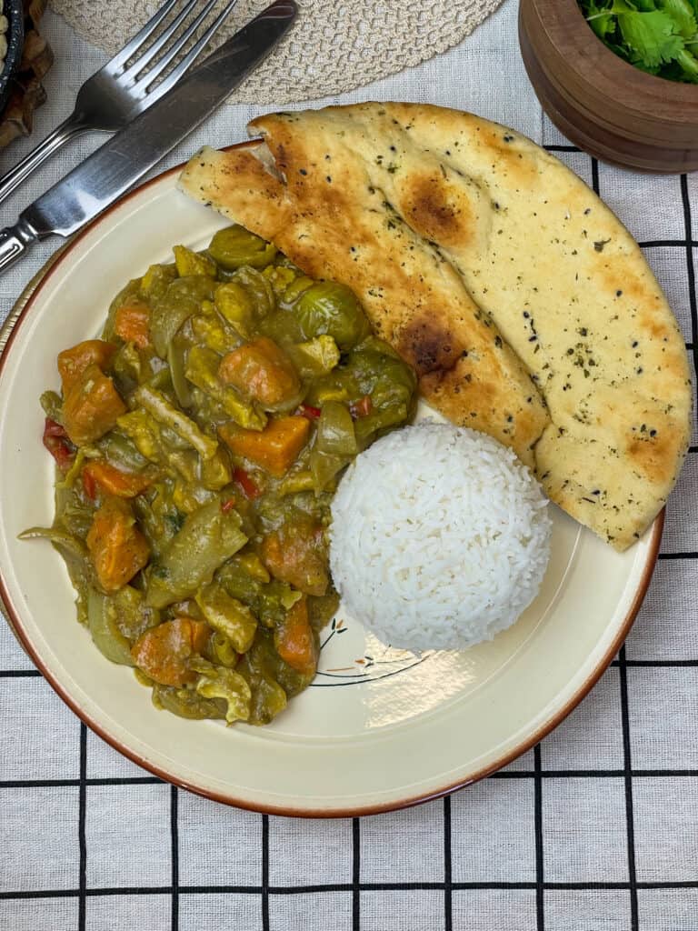 plate of vegan chicken curry with a mound of white rice, slices of naan bread to side, cutlery to side and brown bowl of coriander to side, black and white check table cloth.