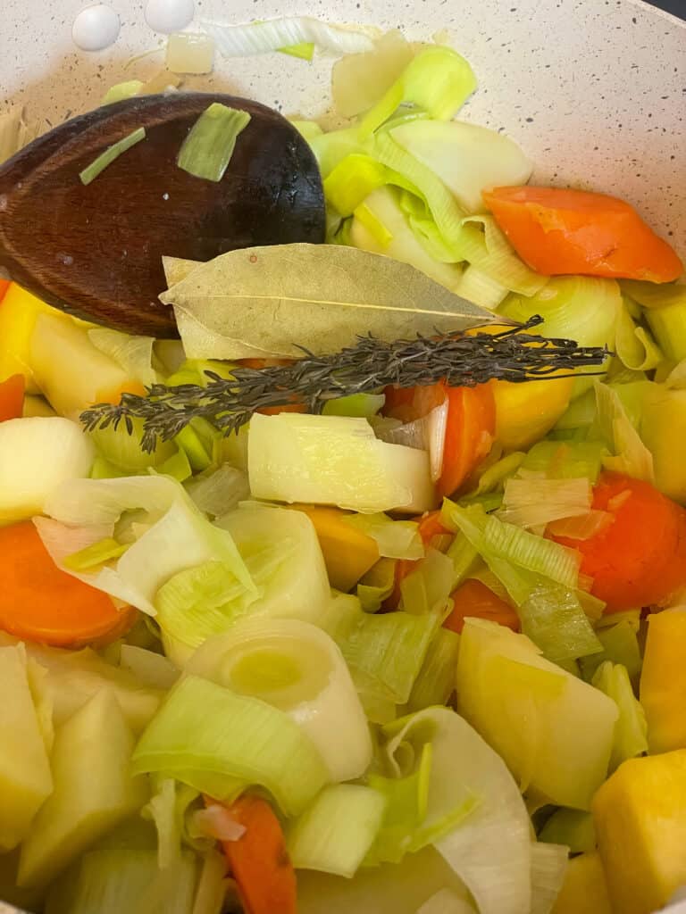 close up of veggies sauteing in pot and a sprig of thyme and bay leaf added, wooden spoon to side.