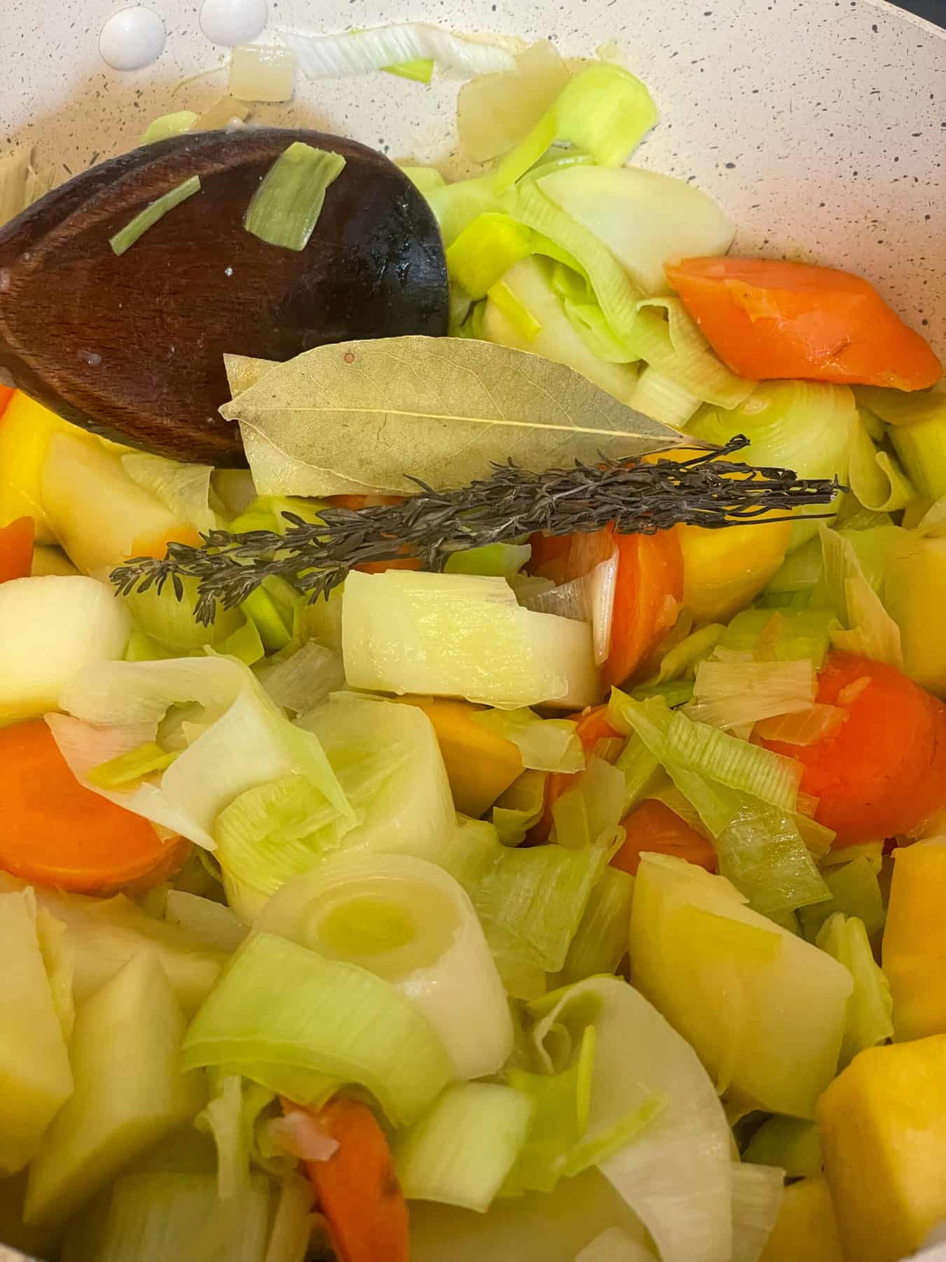 Close up of veggies sauteing in pot and a sprig of thyme and bay leaf added, wooden spoon to side.