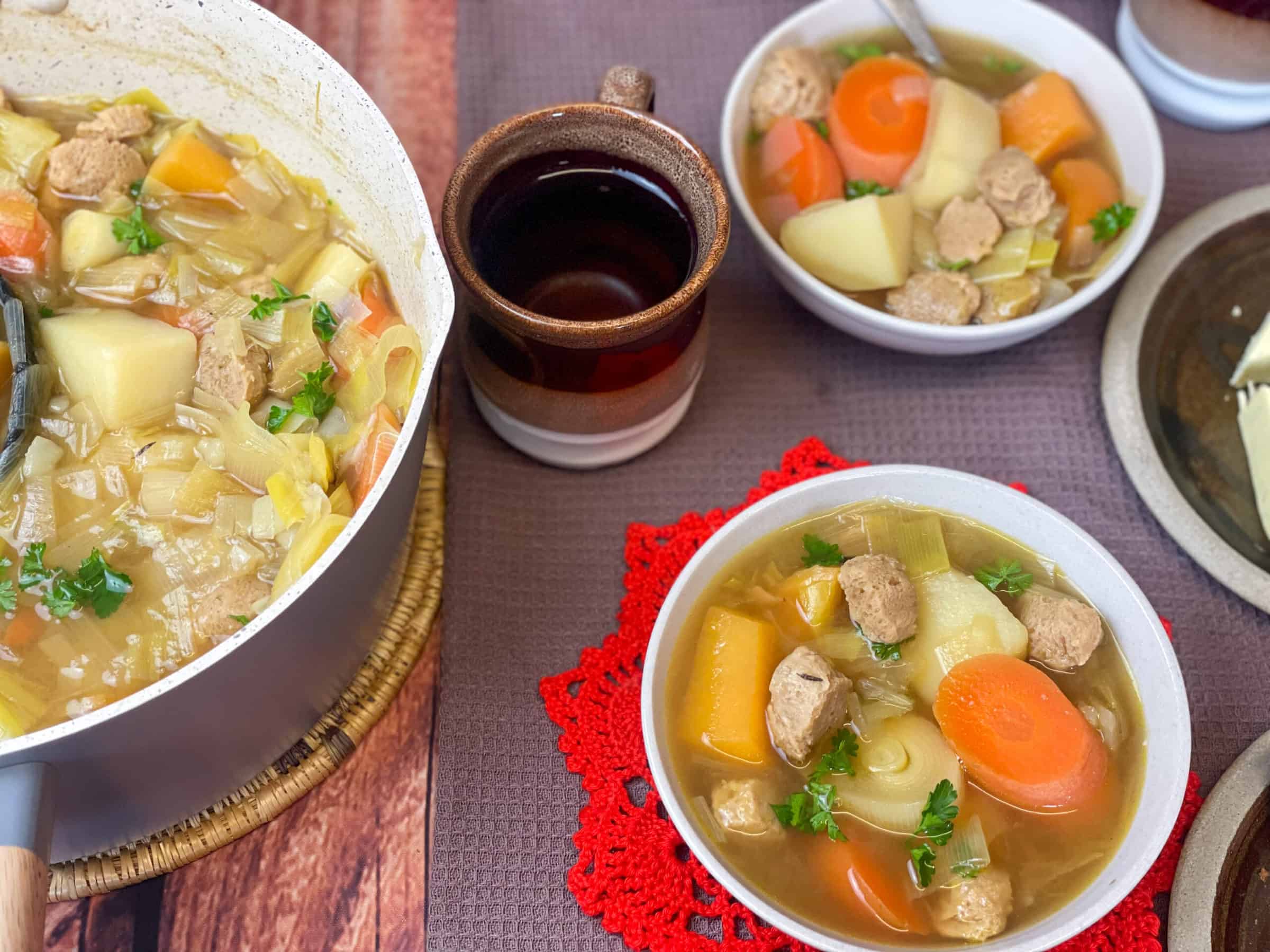 Cooking pot to side with Welsh Cawl soup and two small bowls of soup to side with brown mug, one bowl has a red star shaped crocheted mat, two stone plates to side, brown place mat background.