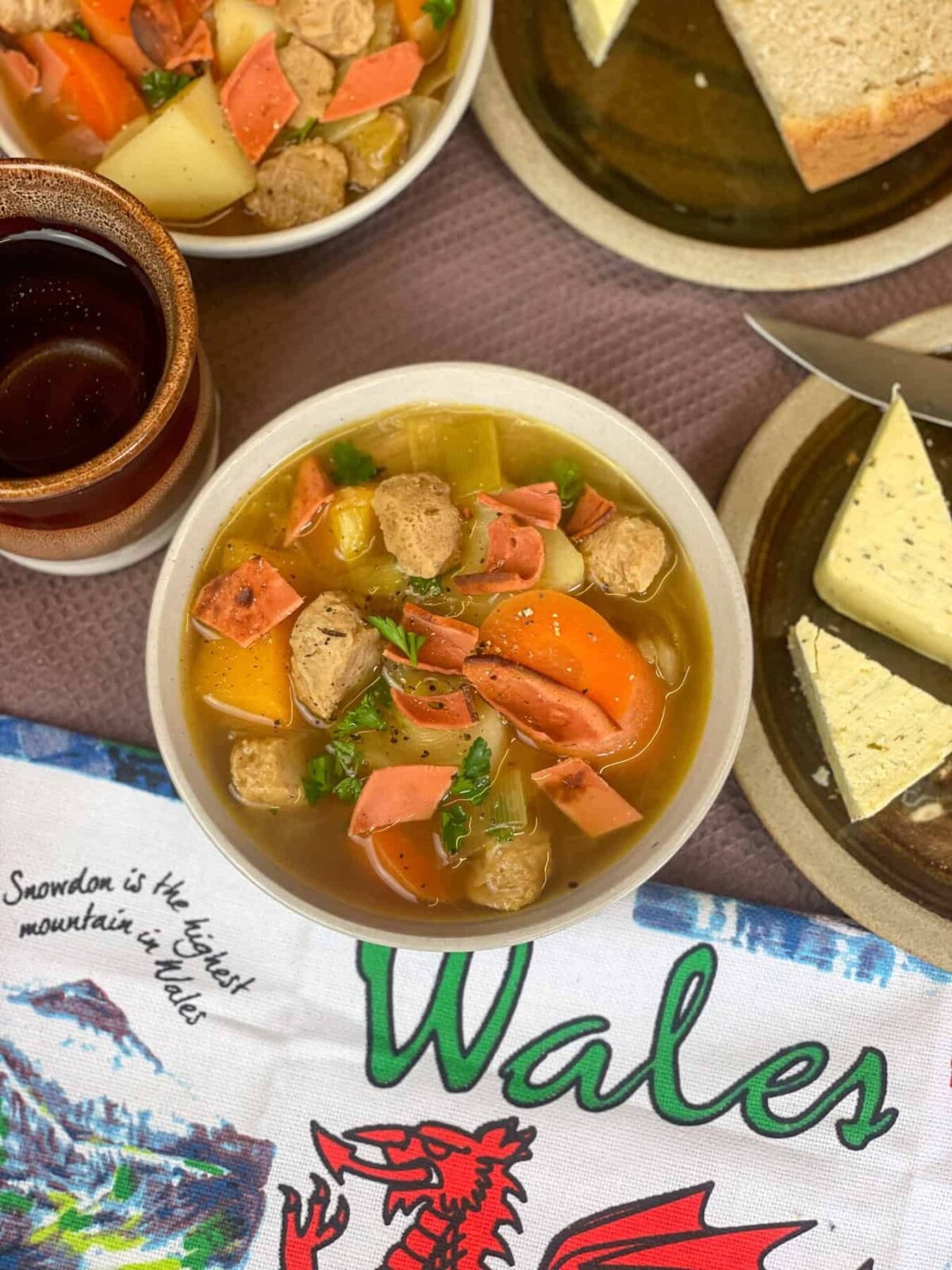Two bowls of vegan Welsh Cawl with small brown plates with bread and cheese to side, brown mugs to side, and Welsh tea towel with the text 'Wales' and a red dragon.