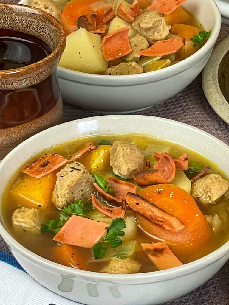 A close up bowl of Welsh Cawl soup in white bowl with second bowl to background, brown and cream mug to side.