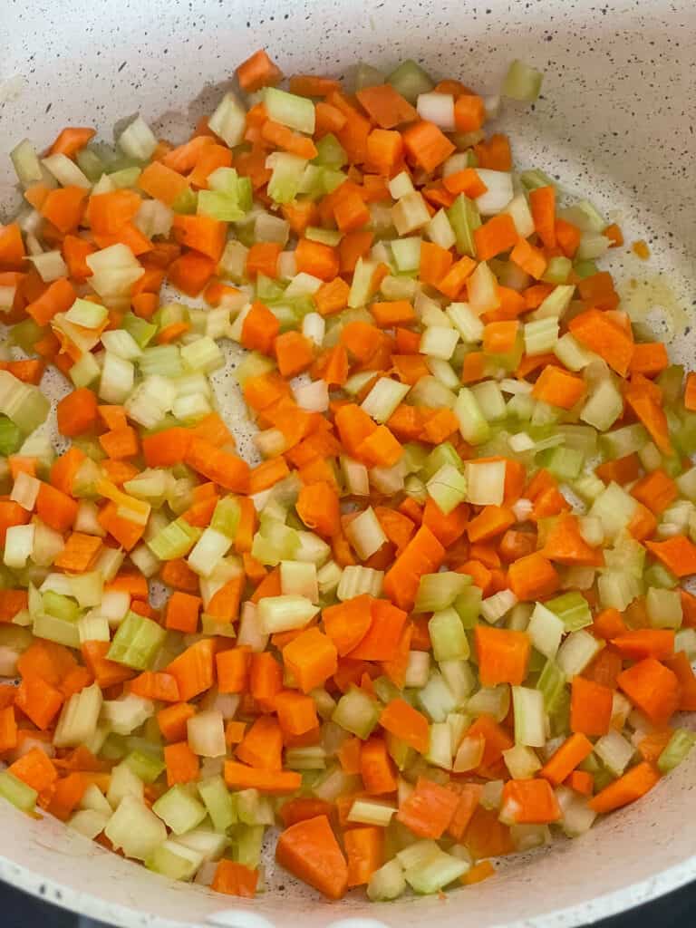 Diced carrot and celery sauteing in cream coloured pot.