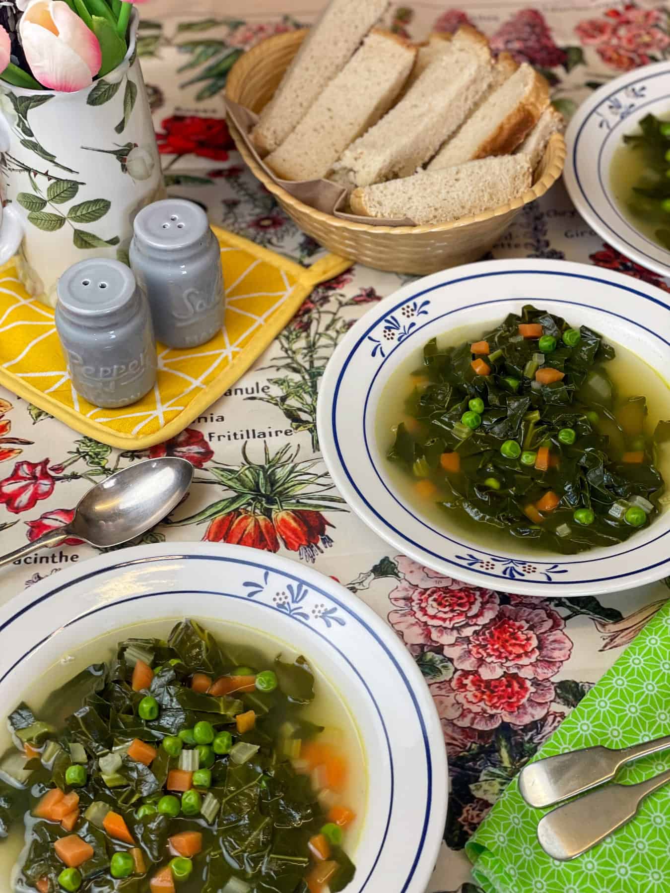 Three bowls of soup served with bread basket, blue salt and pepper shakers, a jug with tulips, and a green and floral background.