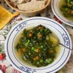 A bowl of spring greens soup with silver spoon, bread basket with bread slices in background, second bowl of soup to side, and colourful floral table cloth.