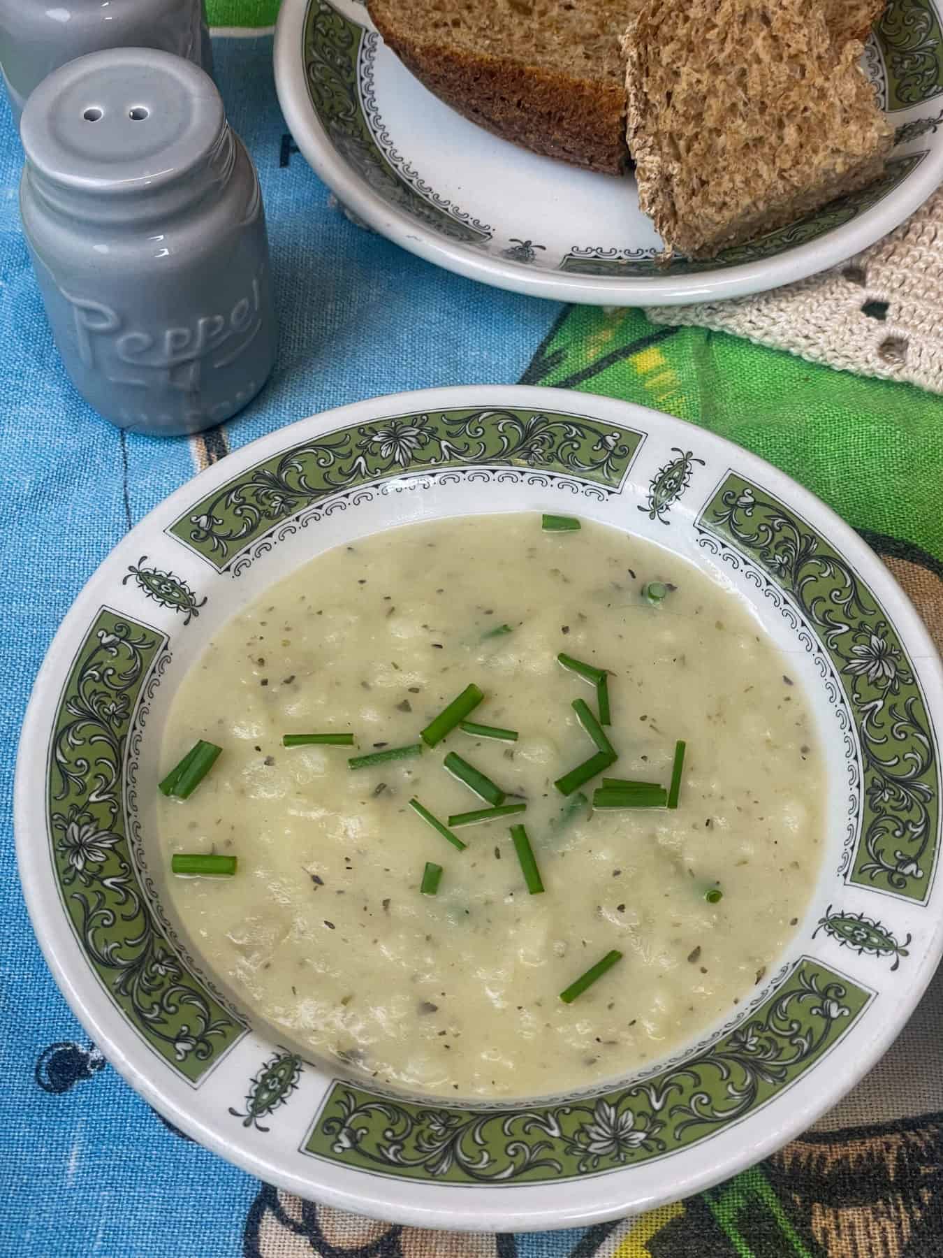 A bowl of Irish potato soup, green pattern on bowl rim, blue background, bread plate to side.