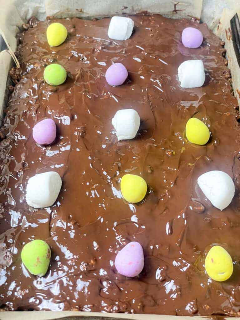 chocolate rice crispy cakes with small mini eggs and marshmallows on top as a decoration, ready to be placed in the refrigerator to set.