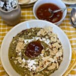 1.Parsnip soup served in white bowl with yellow rim, garnished with mango chutney, toasted sunflower seeds, plant cream and crushed poppadum's. chutney bowl to side, small jug with white flowers, plates to side with seeds and crackers, yellow and white tea towel background.,