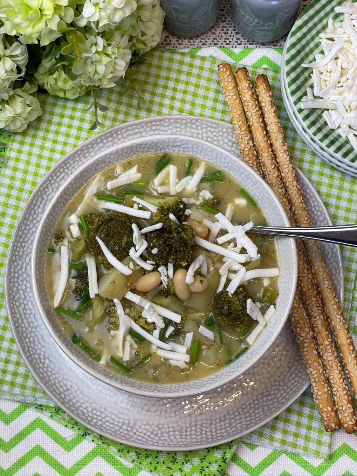A bowl of broccoli cheese soup on plate with three breadsticks, green check plates with shredded cheese to side, green flowers to side, blue salt and pepper pots, green and white layered tablemats.