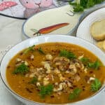 A close up bowl of carrot and cumin soup with walnut and parsley garnish, large spoon with carrot image, mat with rabbit image, and plate to side with crackers, white table cloth background.