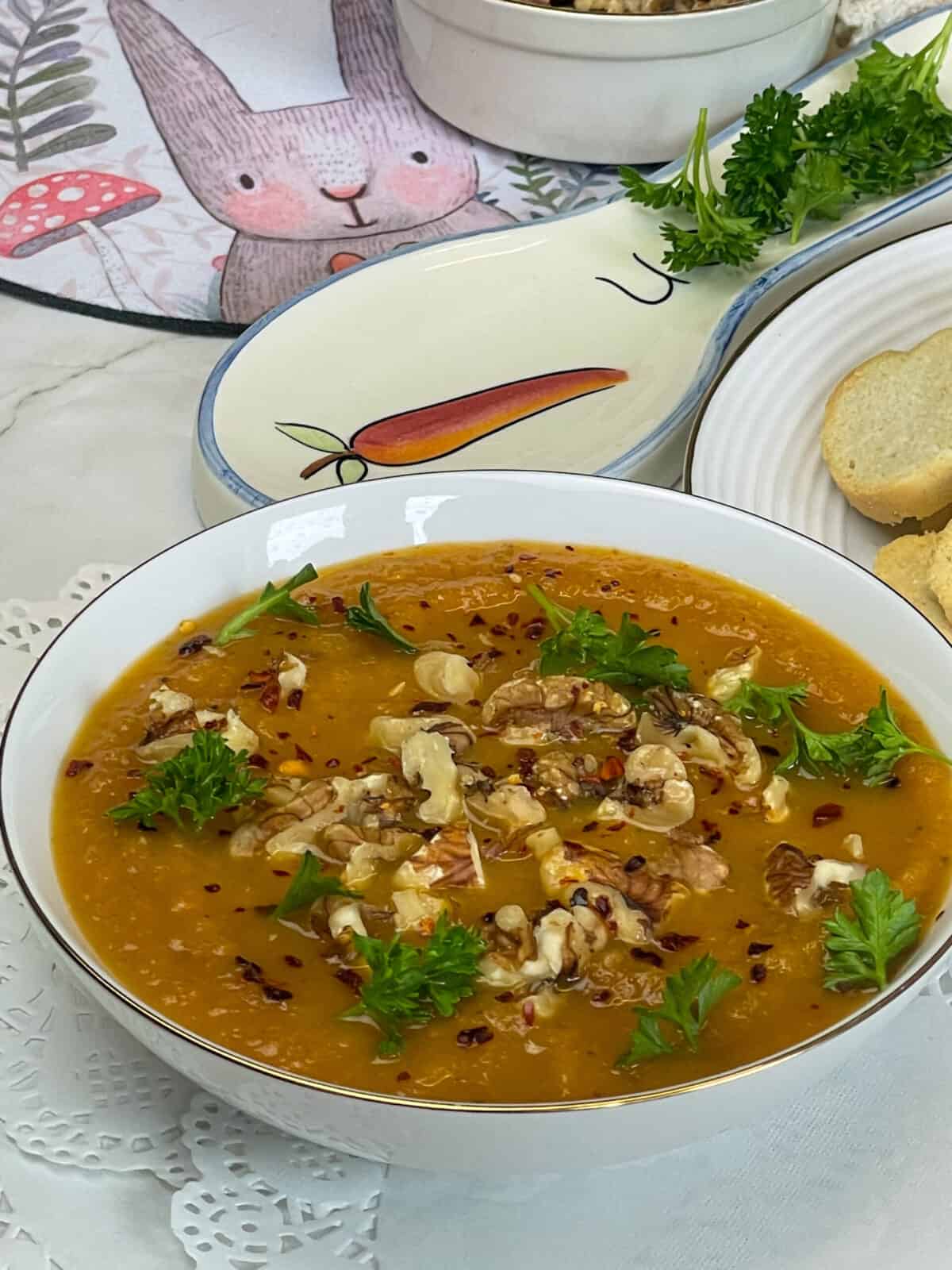 A close up bowl of carrot and cumin soup with walnut and parsley garnish, large spoon with carrot image, mat with rabbit image, and plate to side with crackers, white table cloth background.