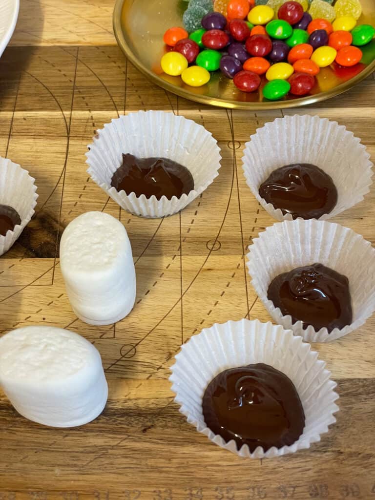 marshmallows on wooden chopping board, melted chocolate in paper cases, gold dish with candy to side.