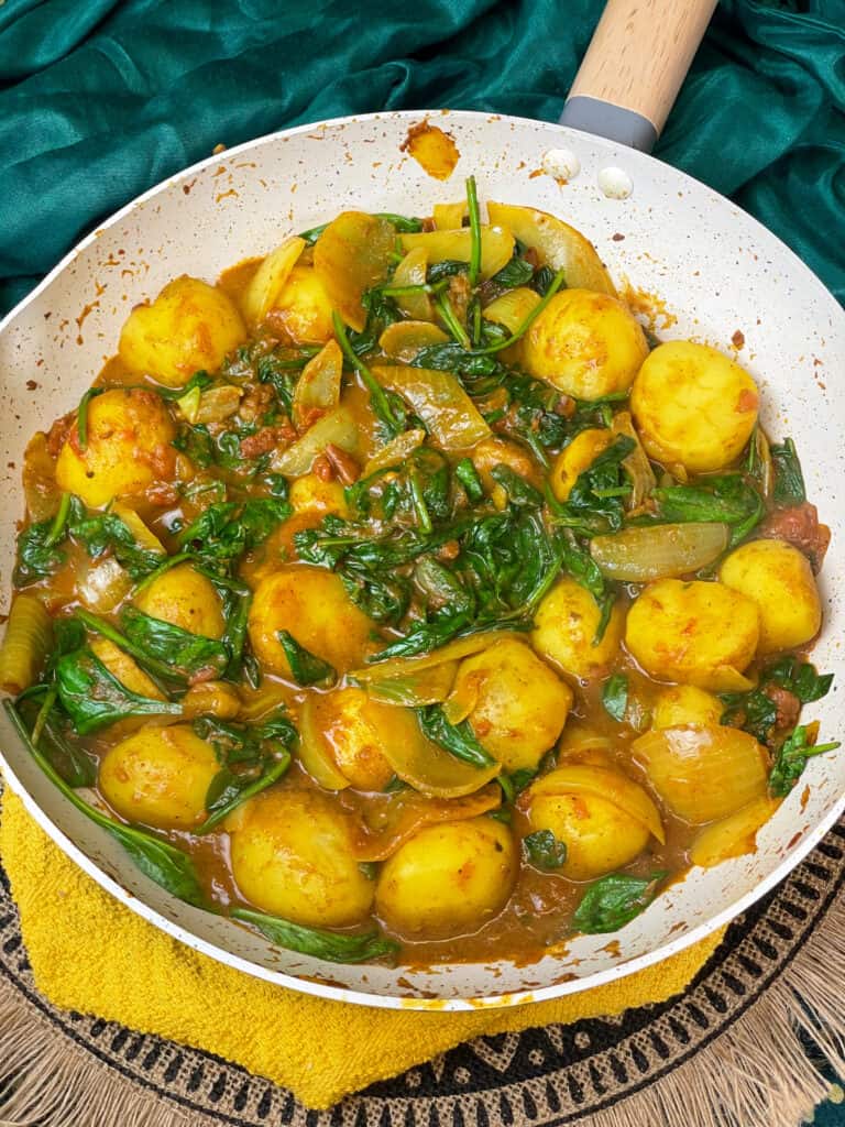 All the spinach mixed through the potatoes and wilted, Saag Aloo ready to serve in pan, with yellow and green background.