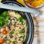 slow cooker pot with rice and peas garnished with fresh herbs, grey ladle across the pot top, yellow, grey and white check background with small spotty bowl filled with tomato flat-bread to side.