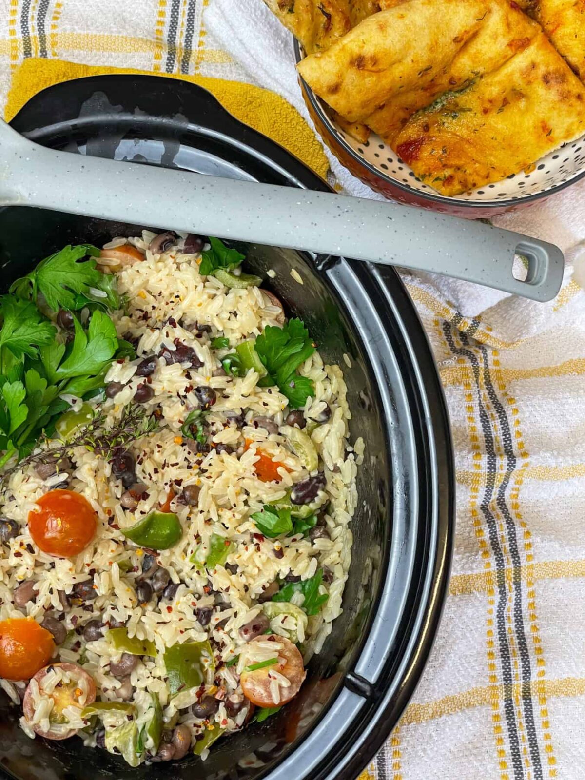 slow cooker pot with rice and peas garnished with fresh herbs, grey ladle across the pot top, yellow, grey and white check background with small spotty bowl filled with tomato flat-bread to side.