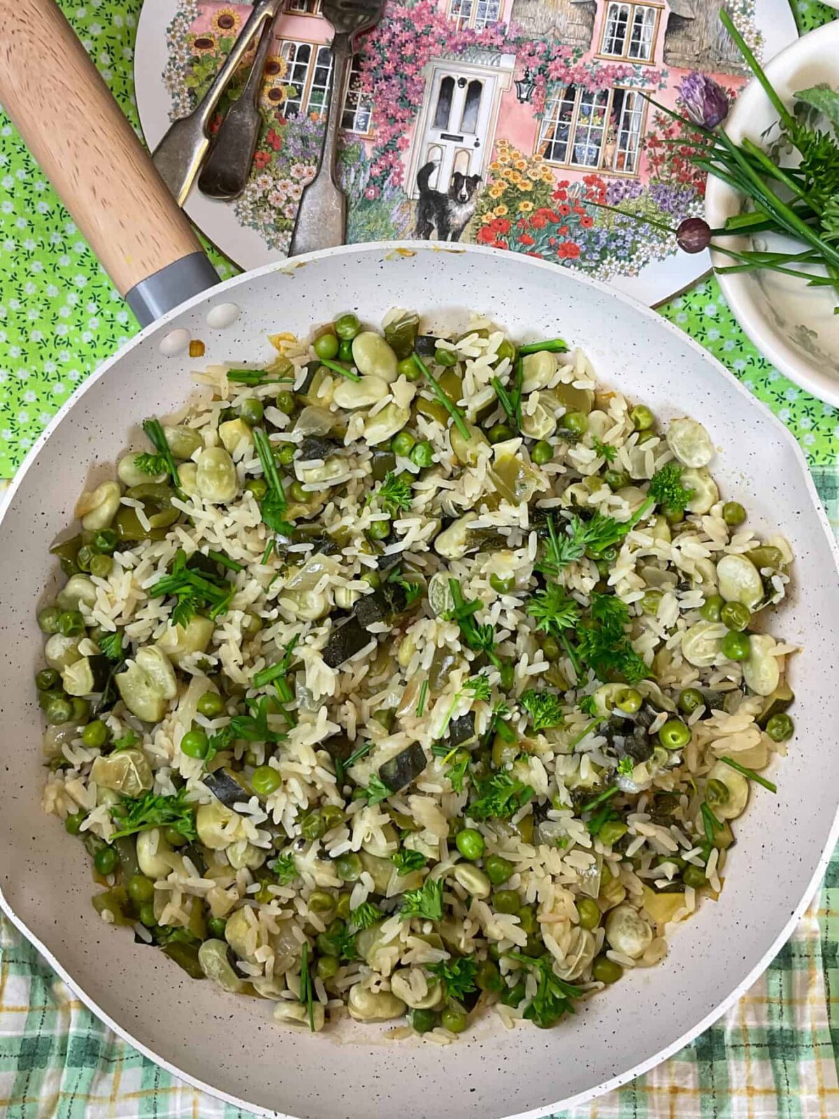 A pan with cooked British pea and broad bean pilaf ready to serve with green and white check tablemat, chopping board to background with cottage and flower image, small dish to side with fresh herbs.