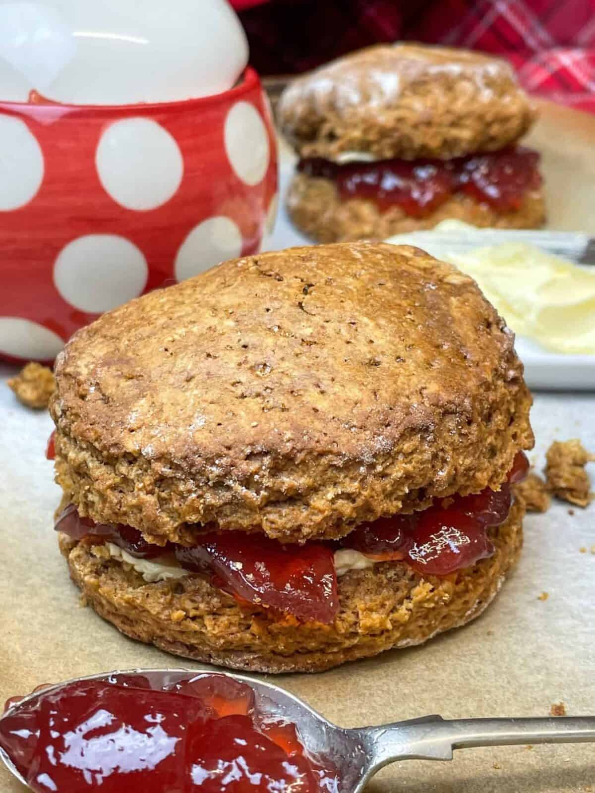 Treacle scone close up, scone filled with strawberry jam and spoon with jam in front, red and white jam pot in background with small white butter dish.