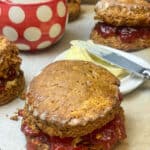 A large scone on baking tray with jam filling, butter dish to background along with red and white spotty jam jar, two treacle scones to side also.