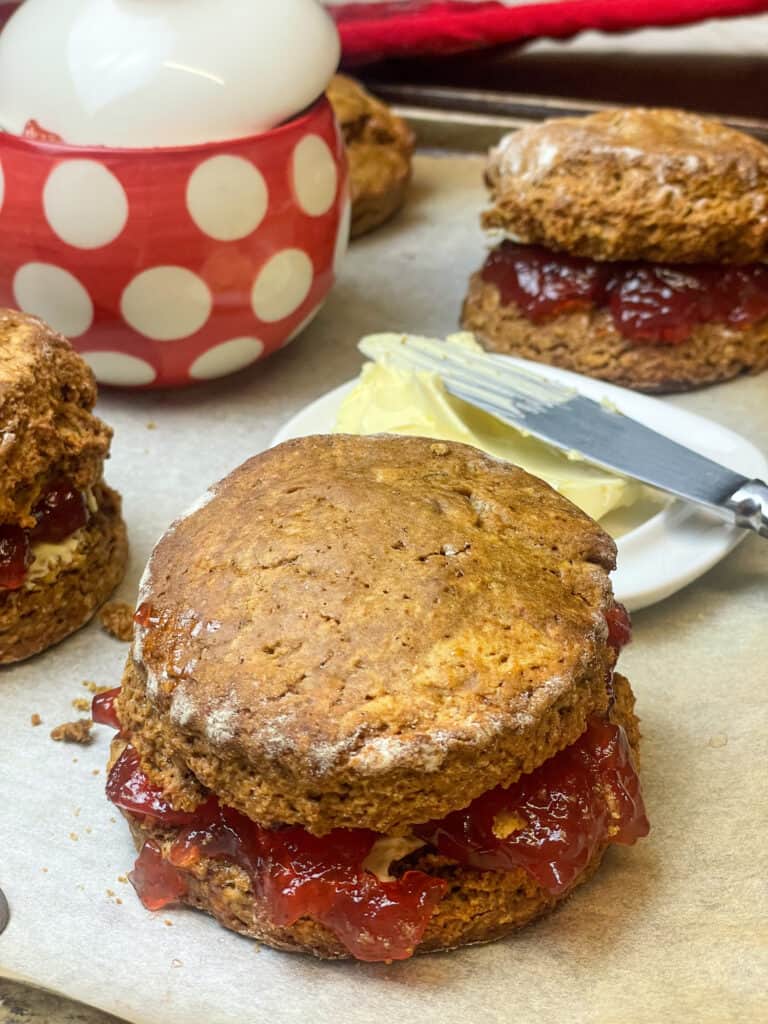A large treacle scone on baking tray with jam filling, butter dish to background along with red and white spotty jam jar, two treacle scones to side also.
