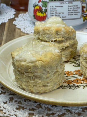 Two cheese scones on a small yellow-rimmed plate with flower image, white doily mat, soup cup with images of tomatoes to distance and more doily's, wooden board background.