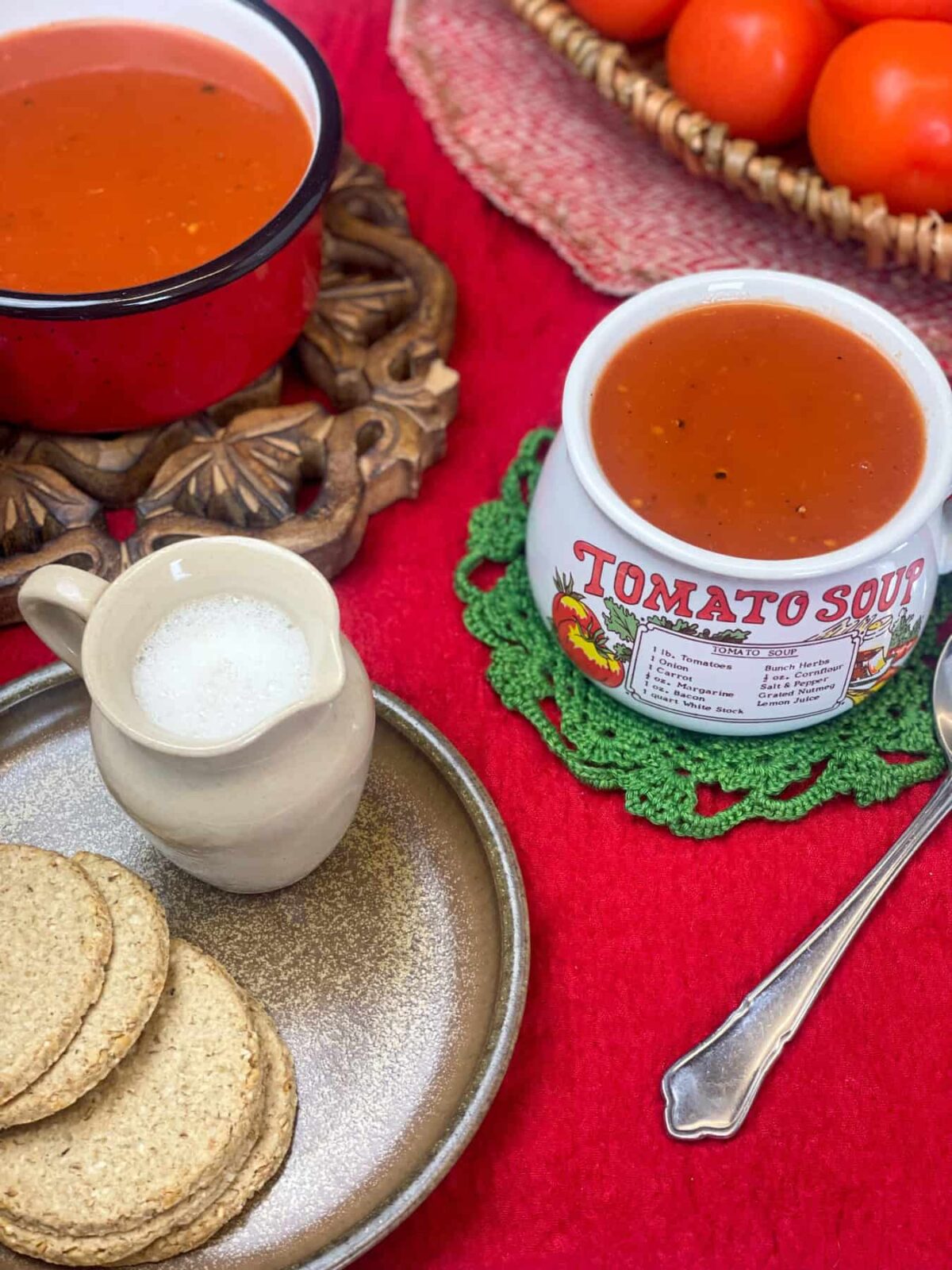 mug of tomato soup on green doily, brown plate with small milk jug and oatcakes, red pot with tomato soup to side on a brown trivet, basket to tomatoes to side, red tablecloth.