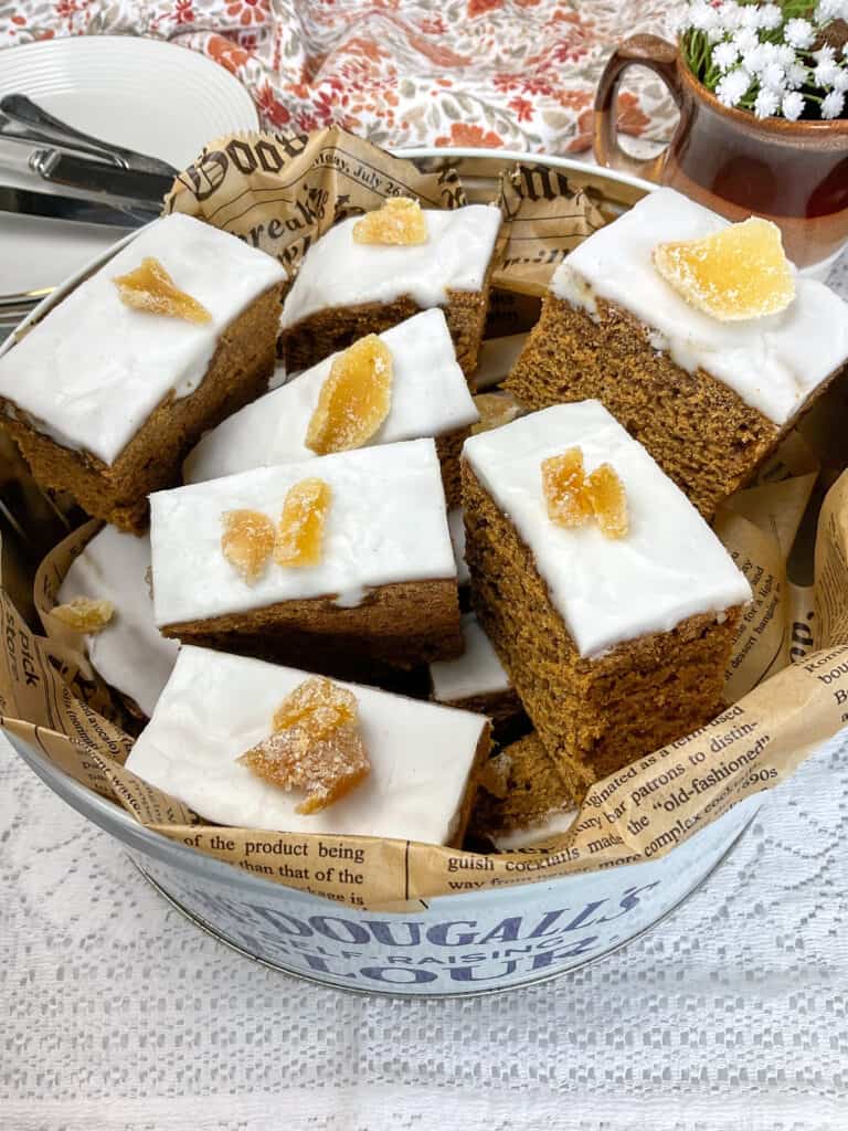 gingerbread sliced into smaller portions and placed into cake tin, white background with brown flower tea towel and mug with white flowers to background.