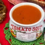 A soup mug with the text 'tomato soup' filled with tomato soup, green doily napkin, red tablecloth, basket of fresh tomatoes to side.