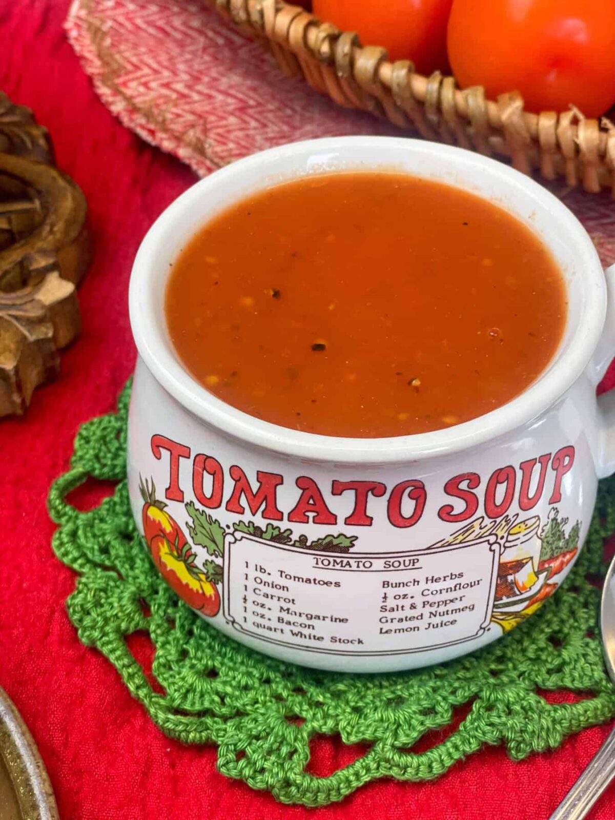 A soup mug with the text 'tomato soup' filled with soup, green doily napkin, red tablecloth, basket of fresh tomatoes to side.