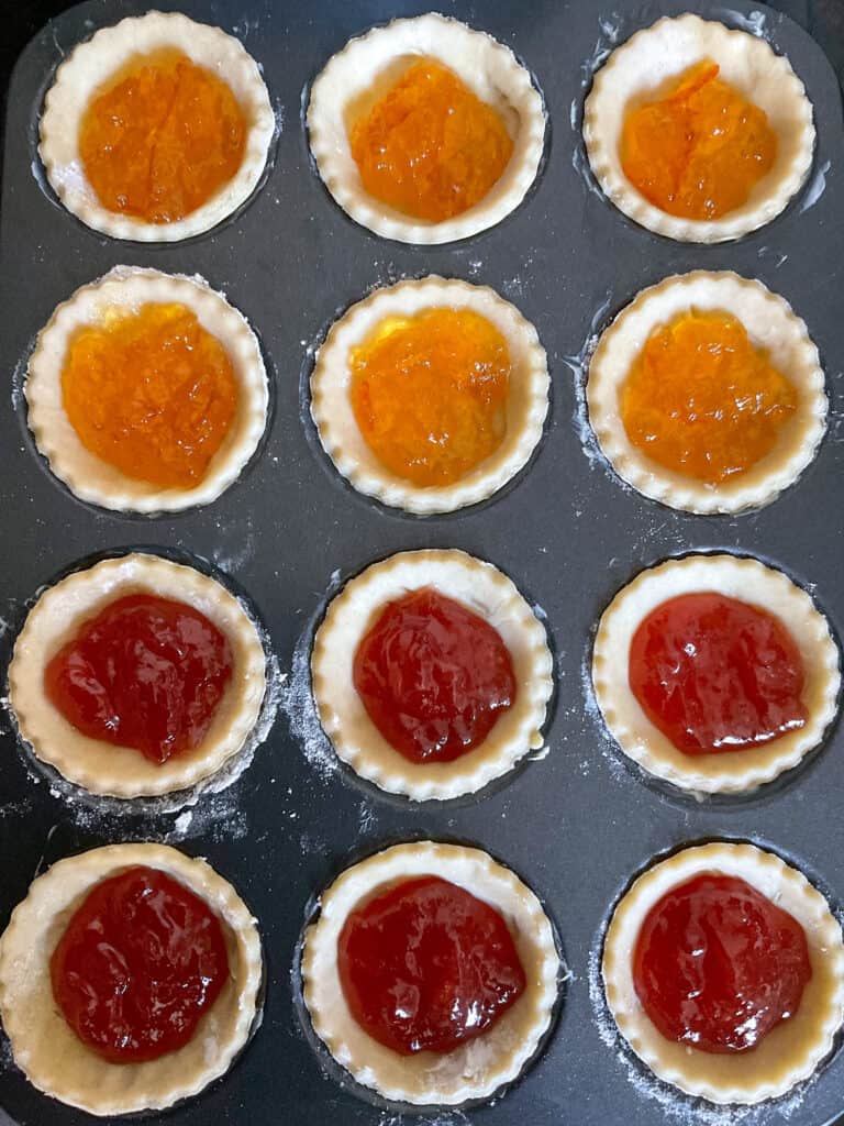 fairy cake tray lined with pastry discs and filled with strawberry jam and marmalade ready to bake in the oven.