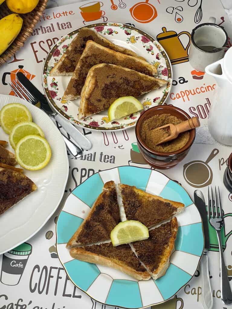 three plates of cinnamon toast with slices of lemon, basket with bananas and a whole lemon to edge of photo, coffee pot to side, small brown bowl of cinnamon sugar with wooden scoop.