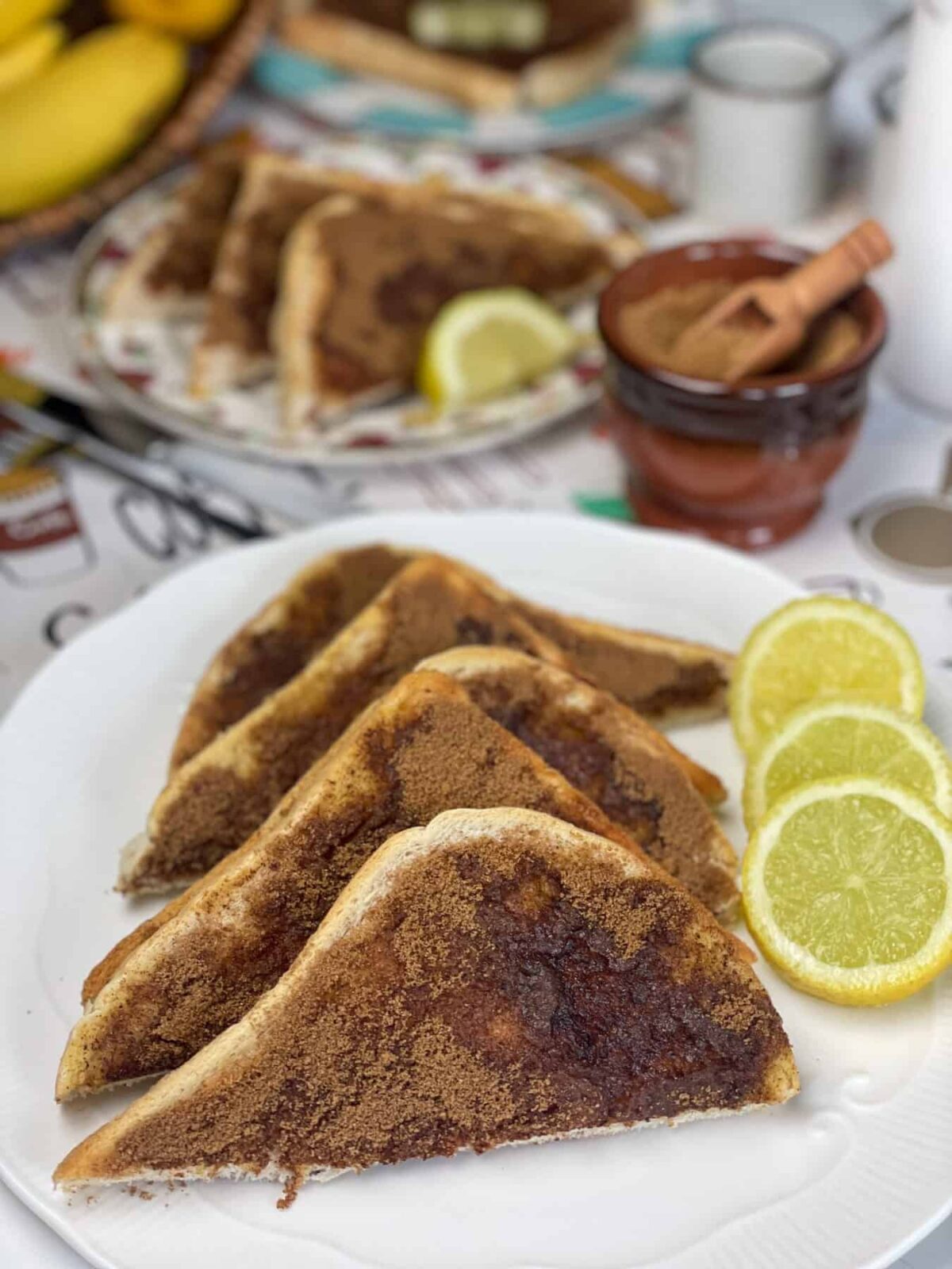 white plate with cinnamon toast and three slices of lemon, extra plates with cinnamon toast to background,coffee pot and cups to background.