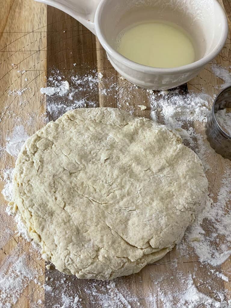 buttermilk scone mixture patted into a dough and ready to be stamped out with a biscuit cutter, white measuring jug to side with buttermilk and wooden chopping board background.