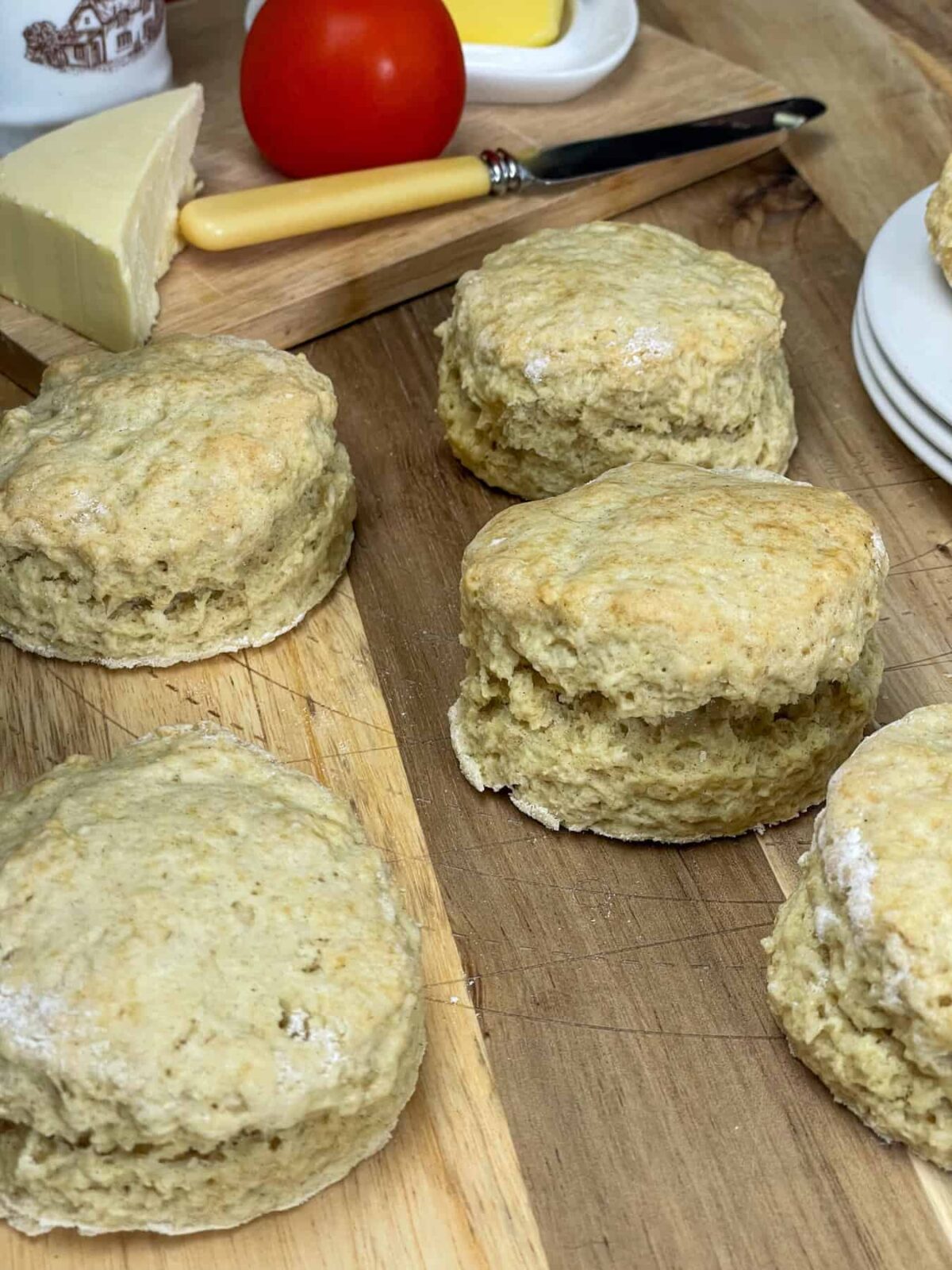 wooden chopping board with five buttermilk scones, small wooden board to background with one tomato, small butter dish, butter knife, wedge of vegan cheese and small pot of chutney.