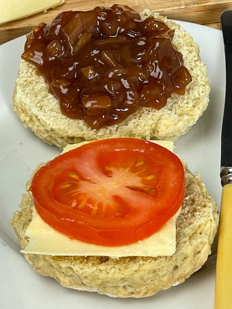 A split scone filled with chutney, sliced tomato and vegan cheese on a white plate.