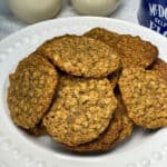 a white plate filled with oatmeal cookies, blue cookie tin to side, two glasses of oat milk to background.