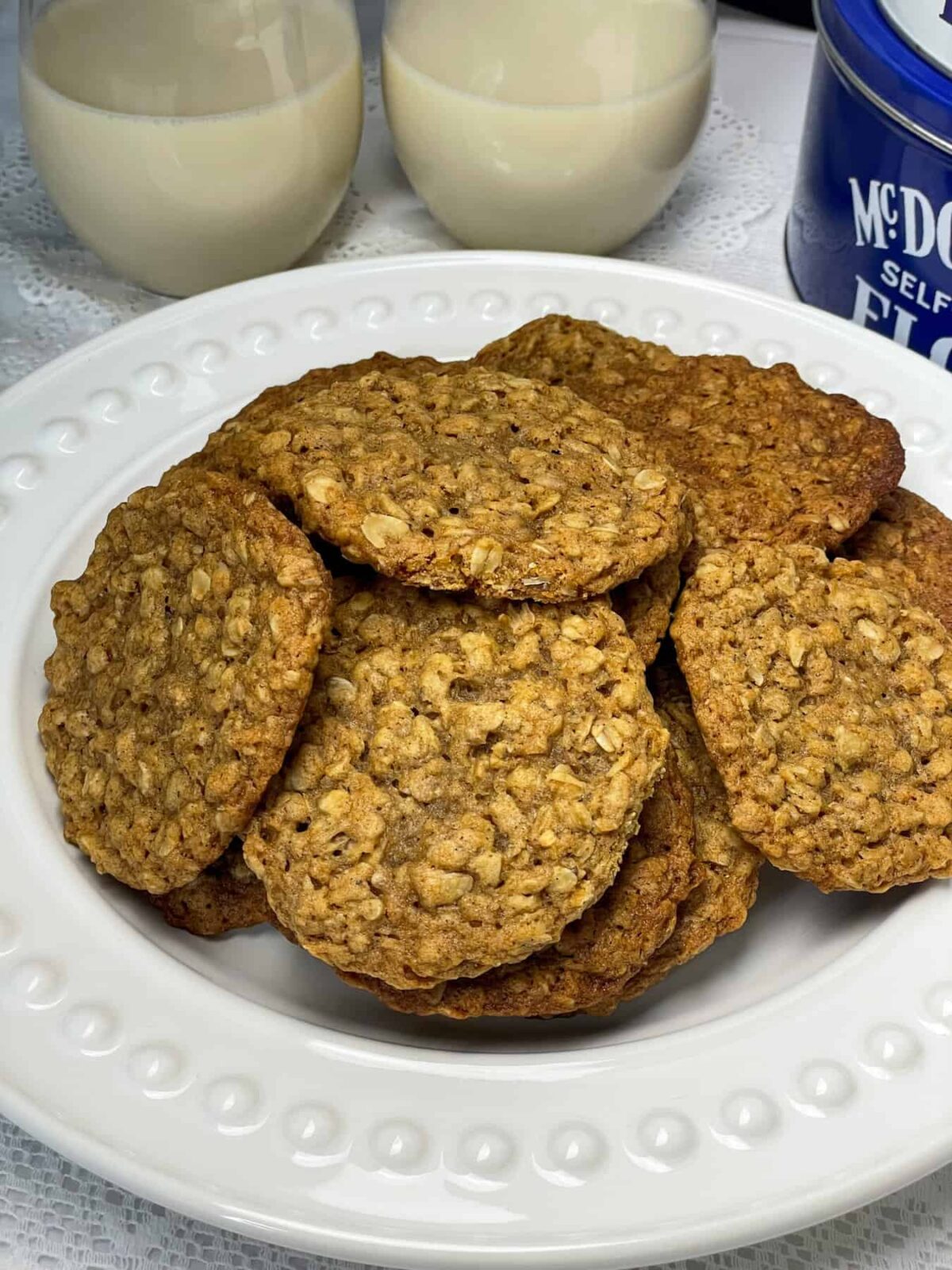 a white plate filled with oatmeal cookies, blue cookie tin to side, two glasses of oat milk to background.