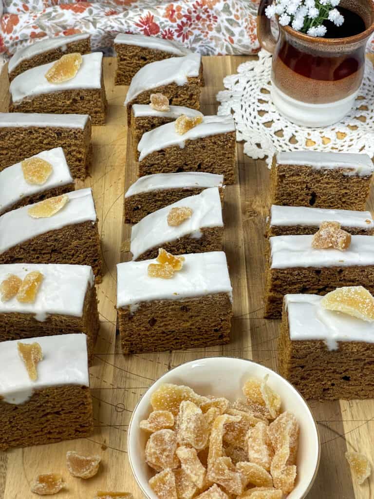 eighteen slices of gingerbread cake on large wooden board, with small dish of crystalized ginger to front, and mug with white flowers to back and brown flower tea towel background.