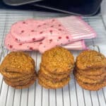 three piles of oatmeal raisin cookies on wire rack with two pink flower oven mitts in background, with pile of baking trays and spatula in far background.