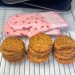 Three piles of oatmeal raisin cookies on wire rack with two pink flower oven mitts in background, with pile of baking trays and spatula in far background.