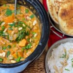 Slow cooker red Thai curry cooked and ready to serve within the slow cooker pot with ladle, to side is a Balti dish with naan breads and a Balti dish with rice garnished with peanuts, red background with cardboard raffia under slow cooker pot.