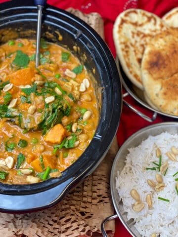 Slow cooker red Thai curry cooked and ready to serve within the slow cooker pot with ladle, to side is a Balti dish with naan breads and a Balti dish with rice garnished with peanuts, red background with cardboard raffia under slow cooker pot.