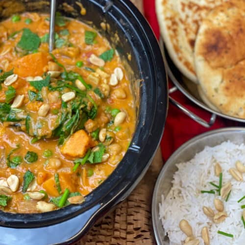 slow cooker red Thai curry cooked and ready to serve within the slow cooker pot with ladle, to side is a Balti dish with naan breads and a Balti dish with rice garnished with peanuts, red background with cardboard raffia under slow cooker pot.