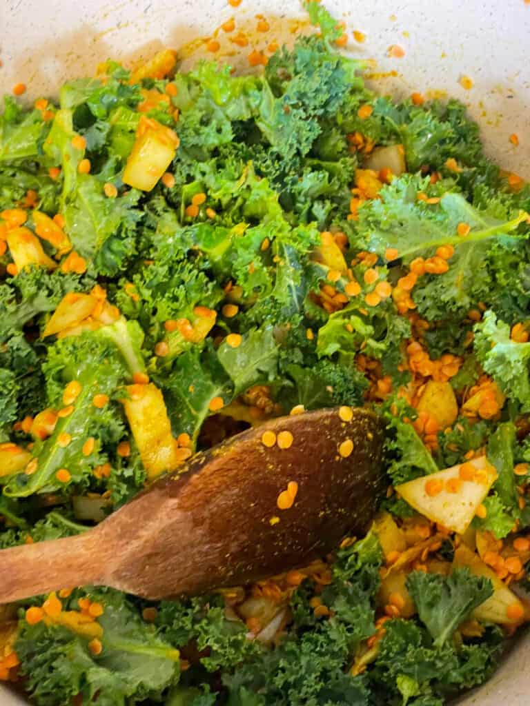 kale added to lentil and ingredients for dahl in the pot with wooden spoon stirring.