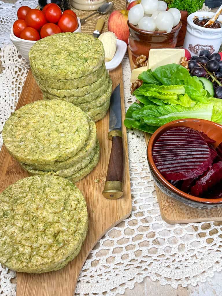 Cutting board with three piles of spinach flavoured rice cakes with brown handled knife to side, and ploughman's lunch platter to side, white doily tablecloth.