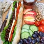 wooden board with vegan ploughman baguette and salad and fruit pieces at the side, small brown dish of pickled onions, small butter dish to back, and cream coloured place mat.