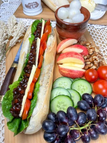 wooden board with vegan ploughman baguette and salad and fruit pieces at the side, small brown dish of pickled onions, small butter dish to back, and cream coloured place mat.