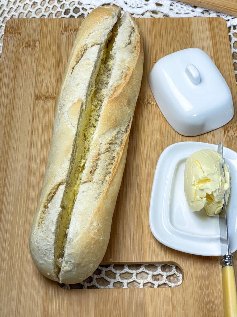 An open baguette on a cutting board with butter dish and knife to side.