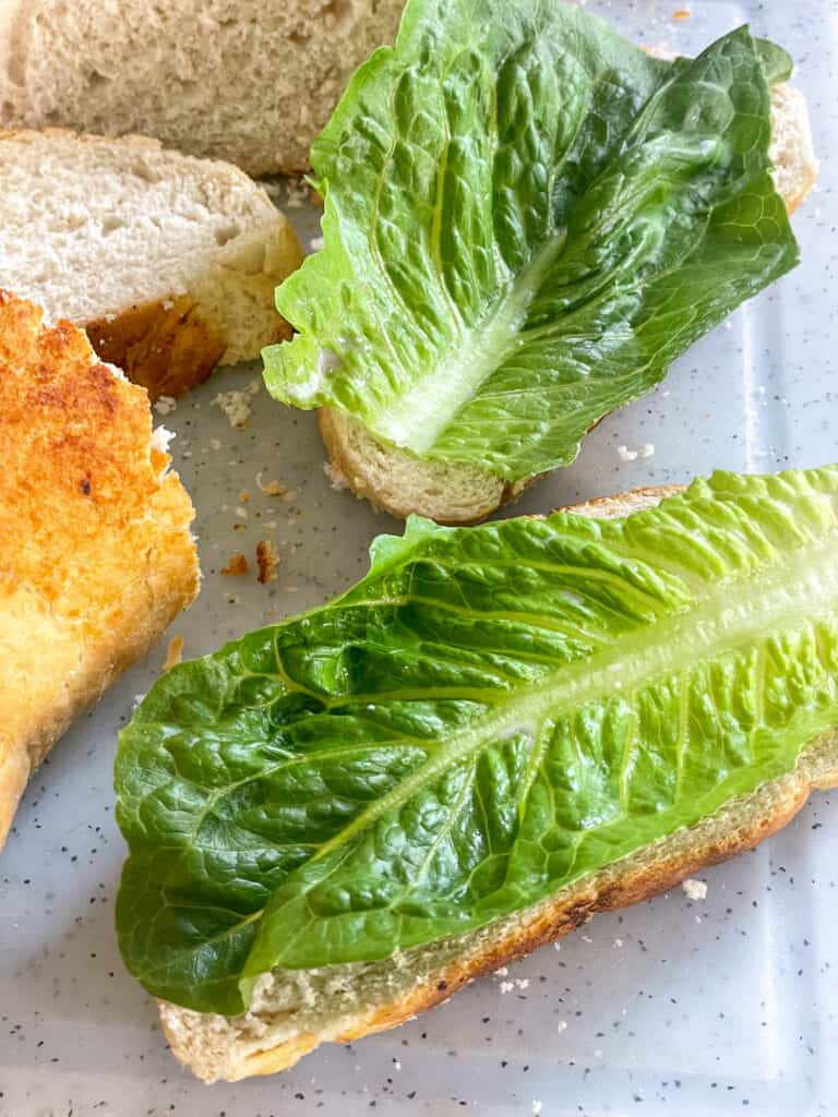 Crusty bread slices with romaine lettuce leaves on top on white chopping board.